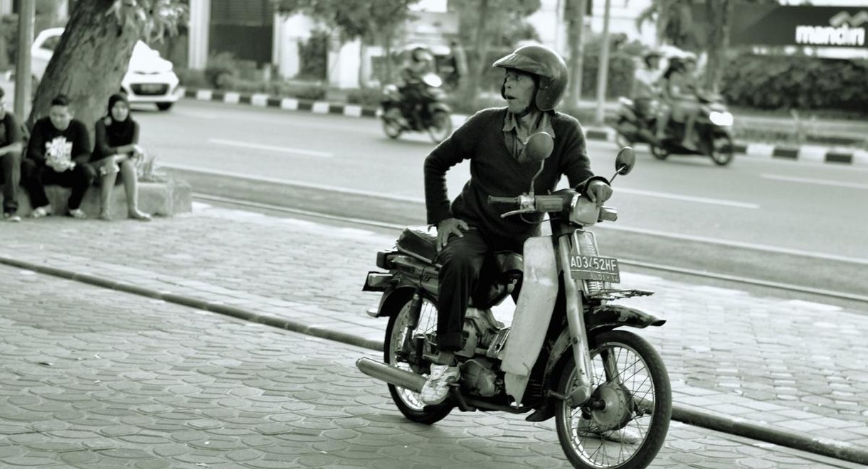 Homme au guidon d'une petite moto ancienne dans une rue, en noir et blanc